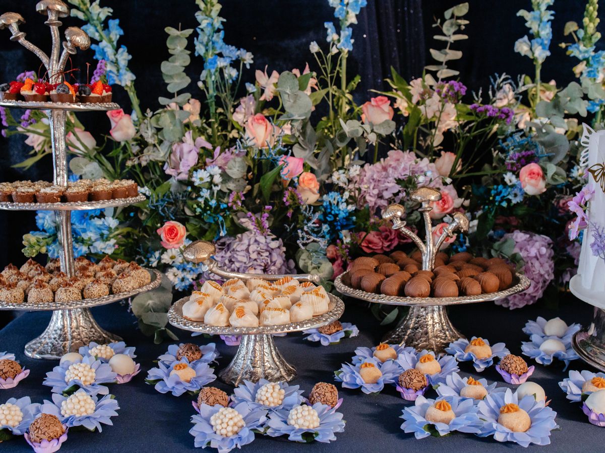 Mesa de doces com arranjos de flores, bandejas de prata e a torre de doces à esquerda