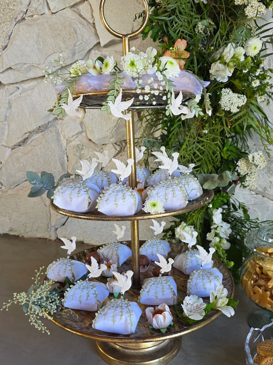 Torre de bandejas com doces azuis e pombas brancas, diante de uma parede de pedra e folhagens