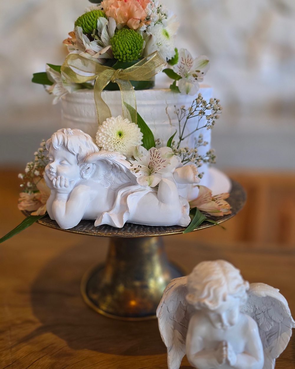 Bolo com flores e um anjinho de gesso deitado sobre uma bandeja dourada, com outro anjo em oração à frente