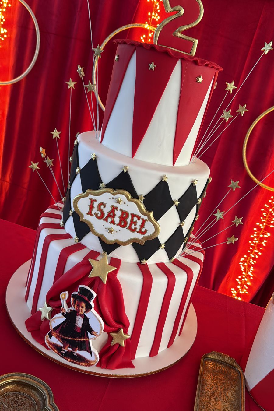 Bolo de três andares com tema de circo, listras vermelhas, losangos pretos, estrelas douradas e o nome Isabel