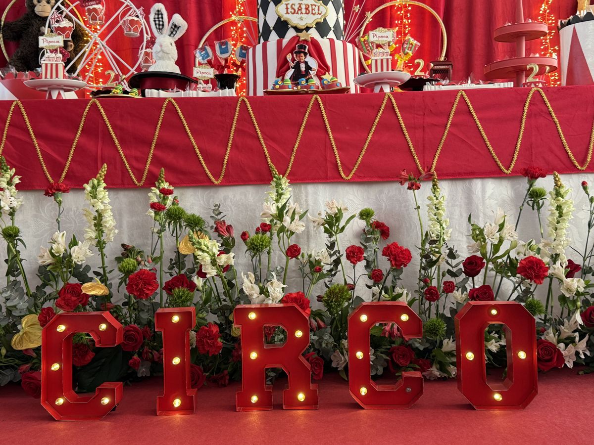 Letras iluminadas formando a palavra CIRCO diante de um arranjo de rosas vermelhas e flores brancas