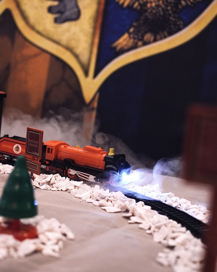 Trem em miniatura do Expresso de Hogwarts sobre trilhos com pedrinhas brancas e fumaça, diante do brasão das casas