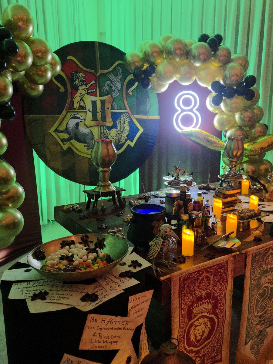 Mesa temática de Harry Potter com brasão de Hogwarts, velas, cartas e arco de balões dourados