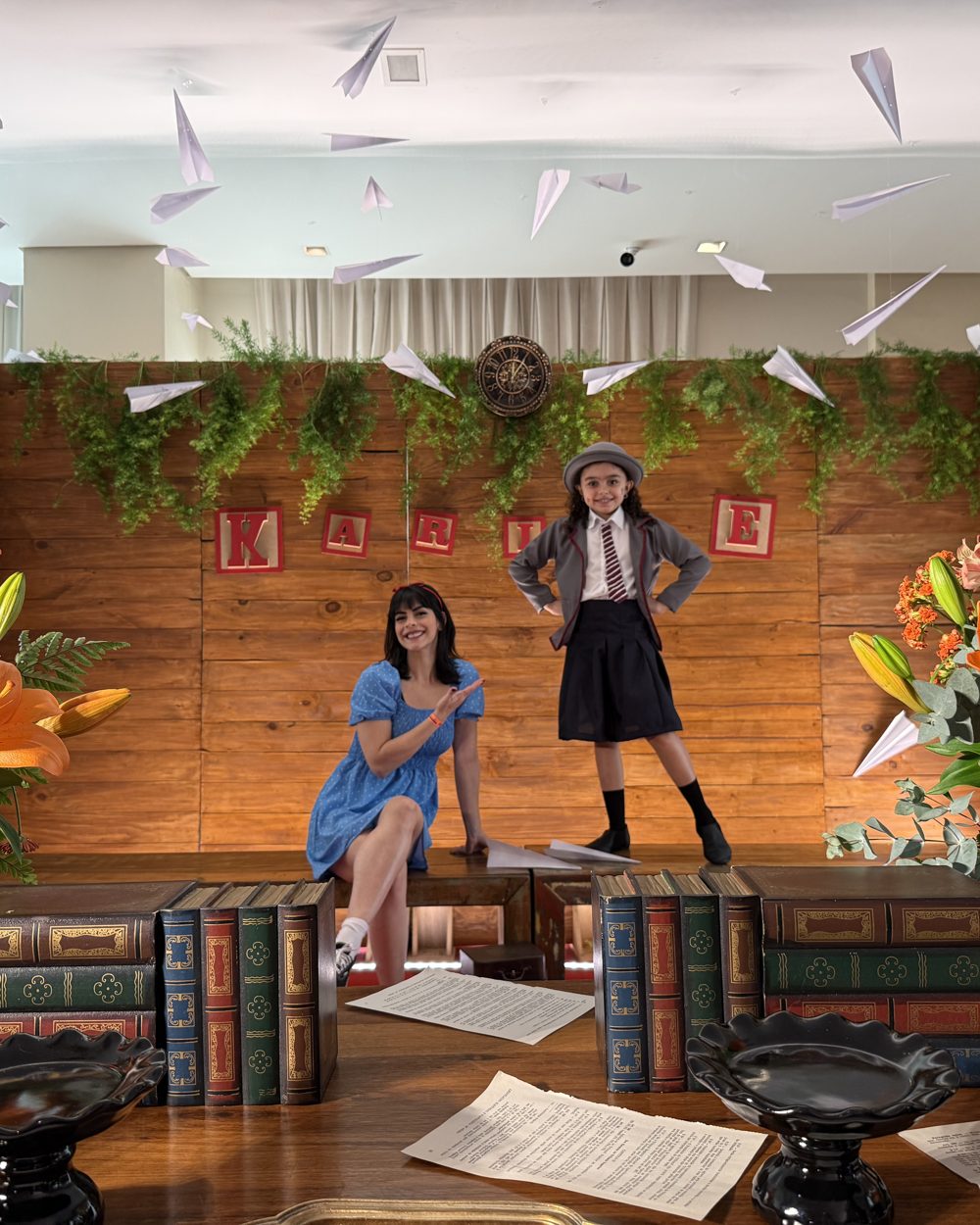 Karine, de uniforme e boina, posa ao lado da Maju sobre o palco da biblioteca da festa temática de Matilda, com aviõezinhos de papel no ar