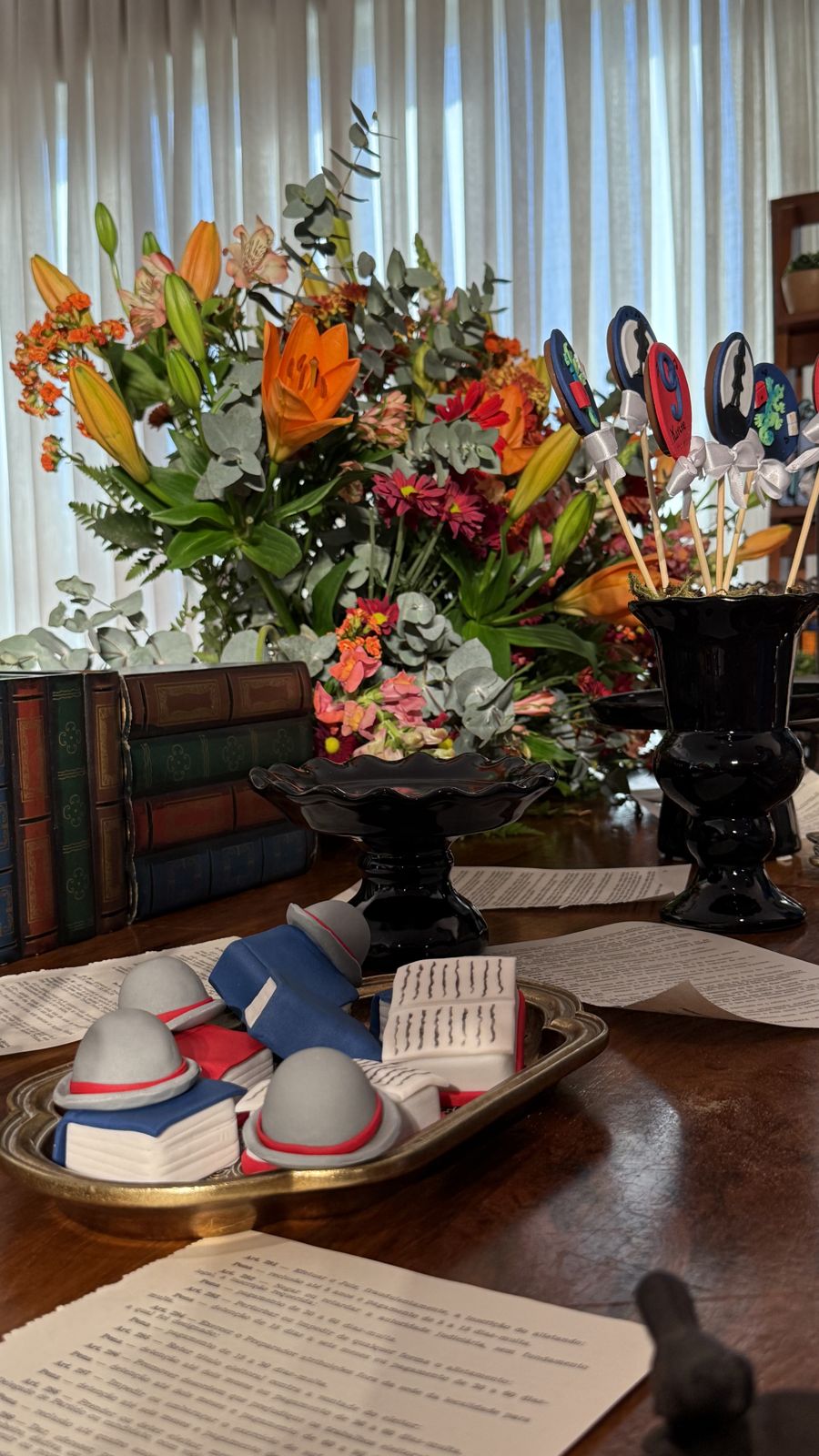 Livrinhos e chapéus numa bandeja dourada, entre flores.