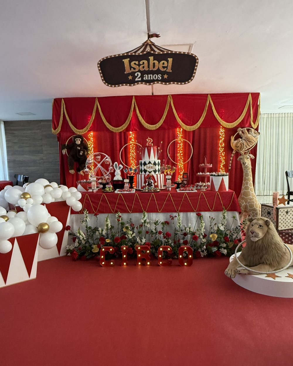 Vista geral da festa de circo da Isabel, com cortina vermelha, letreiro com o nome e a idade dela, girafa, leão e caixas de pipoca feitas de balões